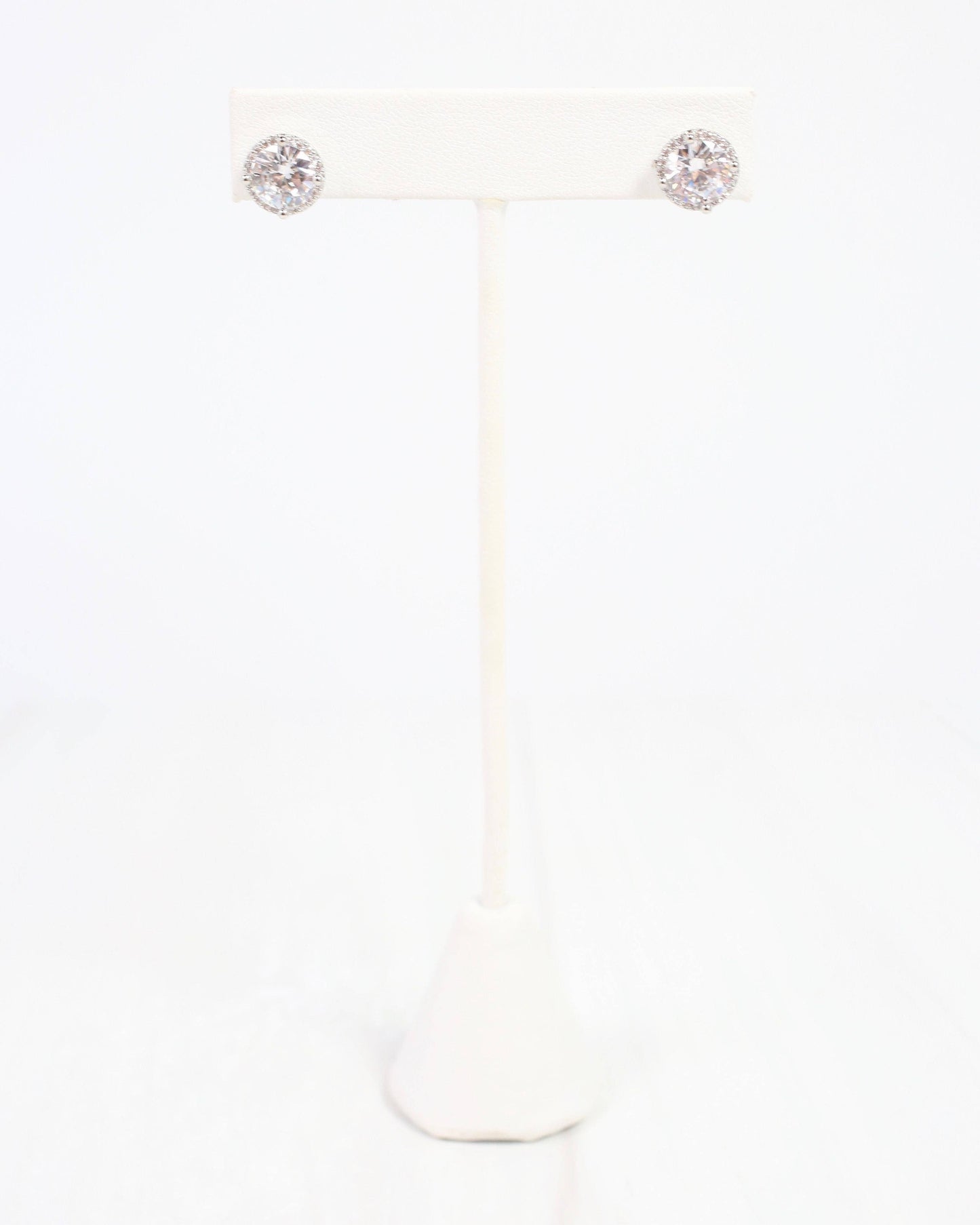 A pair of round stud earrings with sparkling clear gemstones in a silver-toned setting, displayed on a white jewelry stand against a white background.