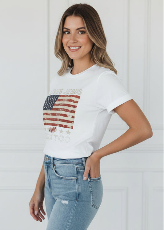 White t-shirt with American flag design and text 'LOVES JESUS AND AMERICA TOO' on a wooden background.