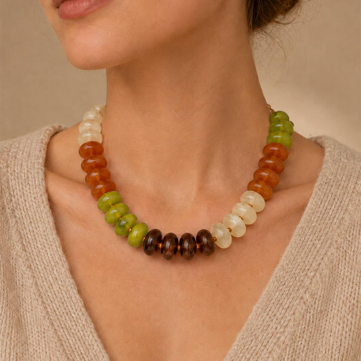A close-up of a woman wearing a beaded necklace made of alternating translucent white, orange-brown, and green beads, with a center section of dark brown beads.