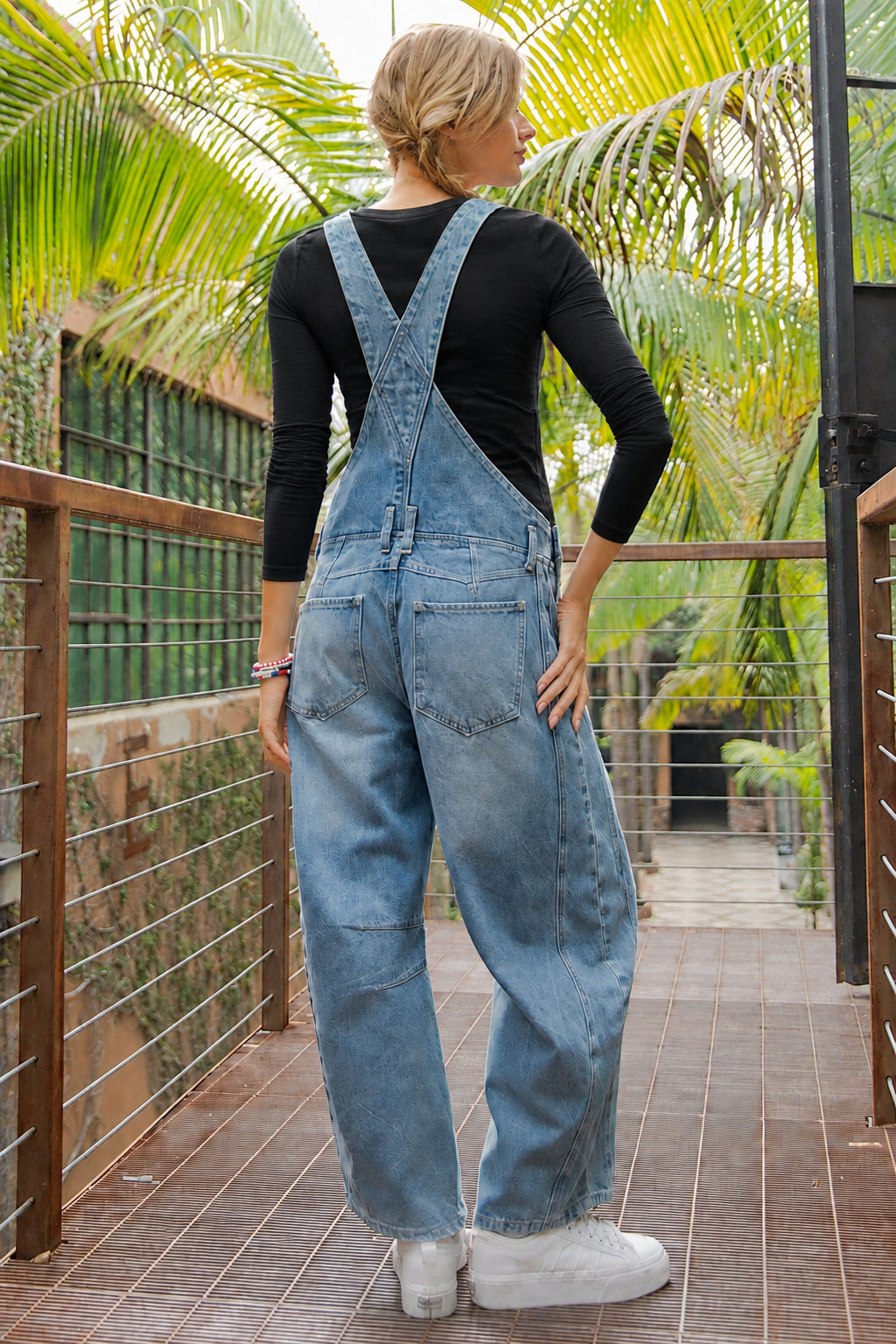 Person wearing blue denim overalls with a red shirt and American flag patch, standing on a wooden deck with greenery in the background.