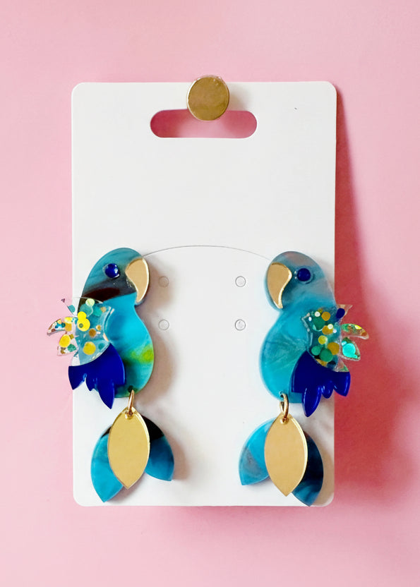 Pair of colorful parrot-shaped earrings on a branded card against a pink background
