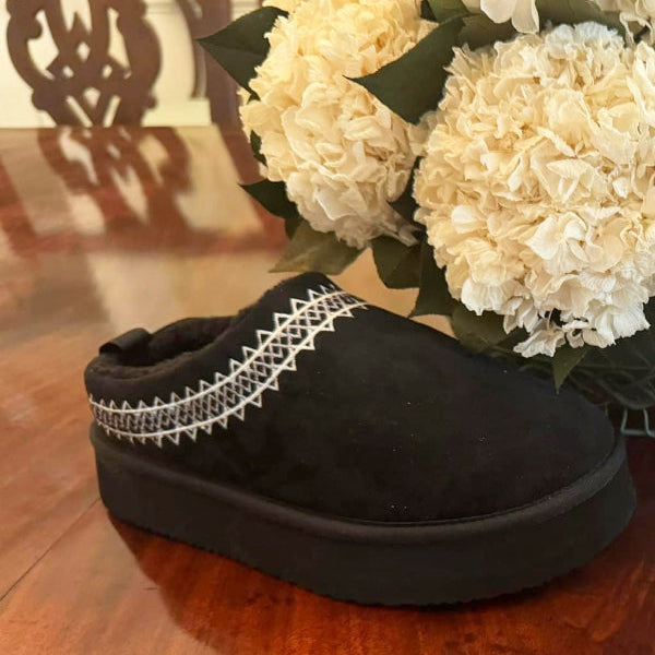 A black faux sherpa slipper with a braided design on the top, placed on a wooden floor next to a bouquet of white flowers.