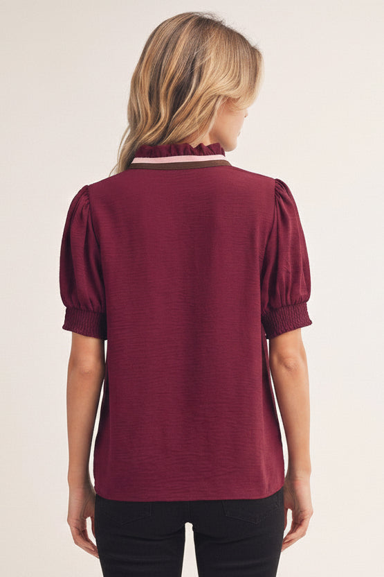 A woman viewed from the back wearing a short-sleeve burgundy top with puff sleeves and a ruffled high collar, paired with white pants, against a plain light beige background.