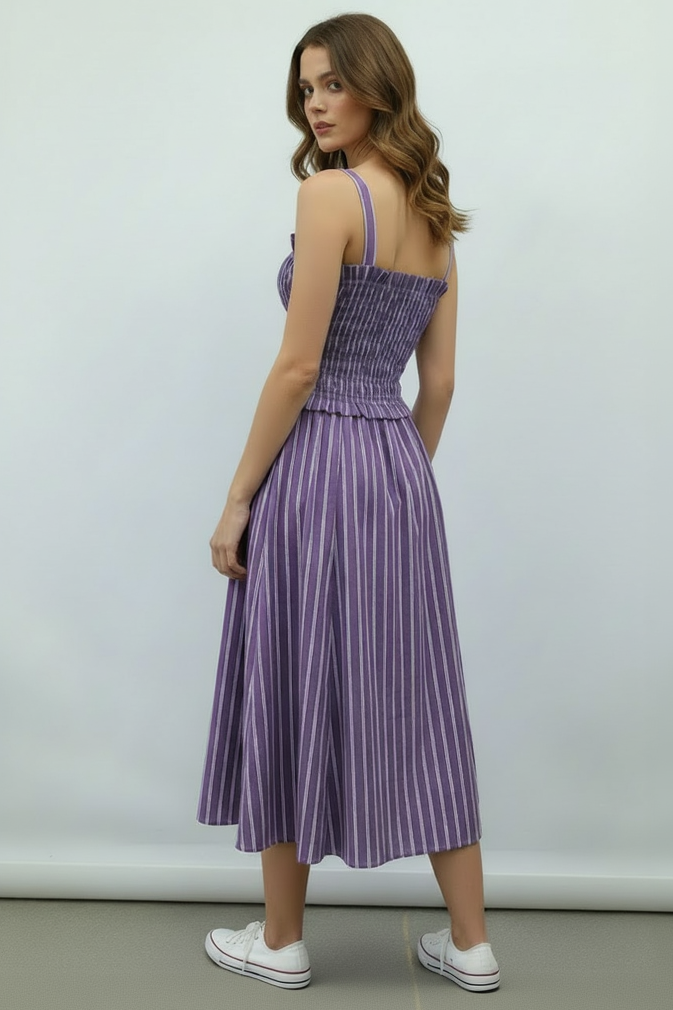 A woman modeling a purple and white vertically striped midi dress with a smocked bodice and thin shoulder straps, shown from a back-three-quarter angle against a plain light gray studio background.
