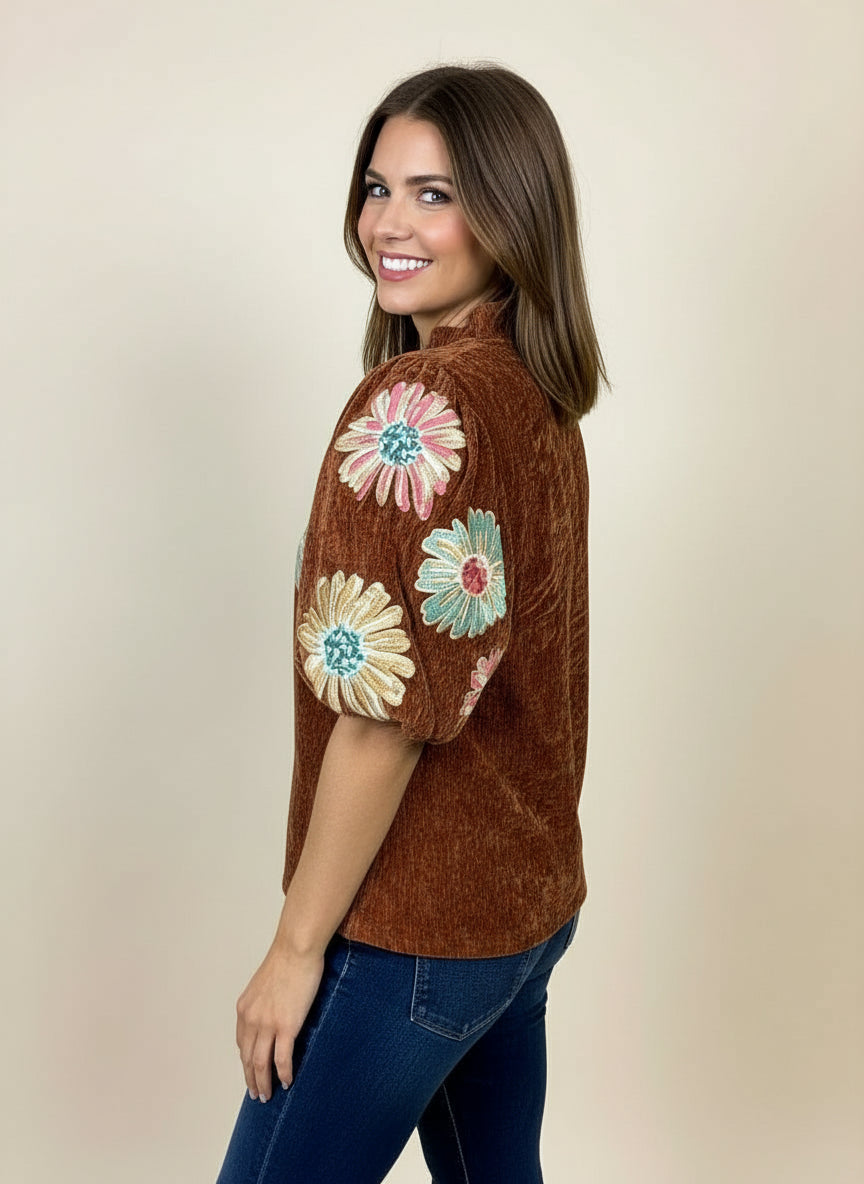 Woman wearing a brown shirt with colorful floral designs on sleeves.