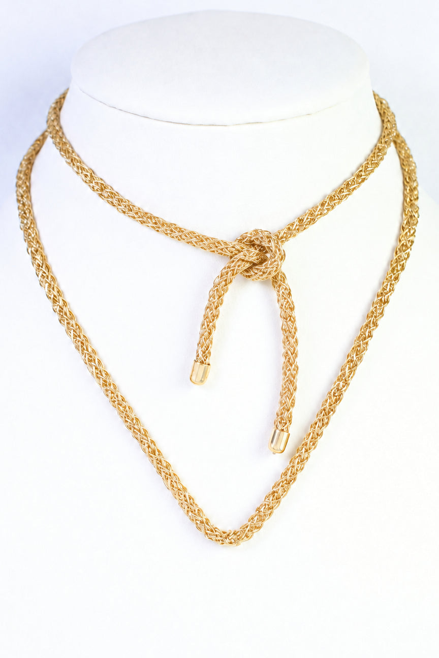 A gold-toned double-layered chain necklace displayed on a white mannequin bust. The top layer is a braided rope-style choker with a tie knot, and the bottom layer is a longer chain ending in a textured cone-shaped pendant.