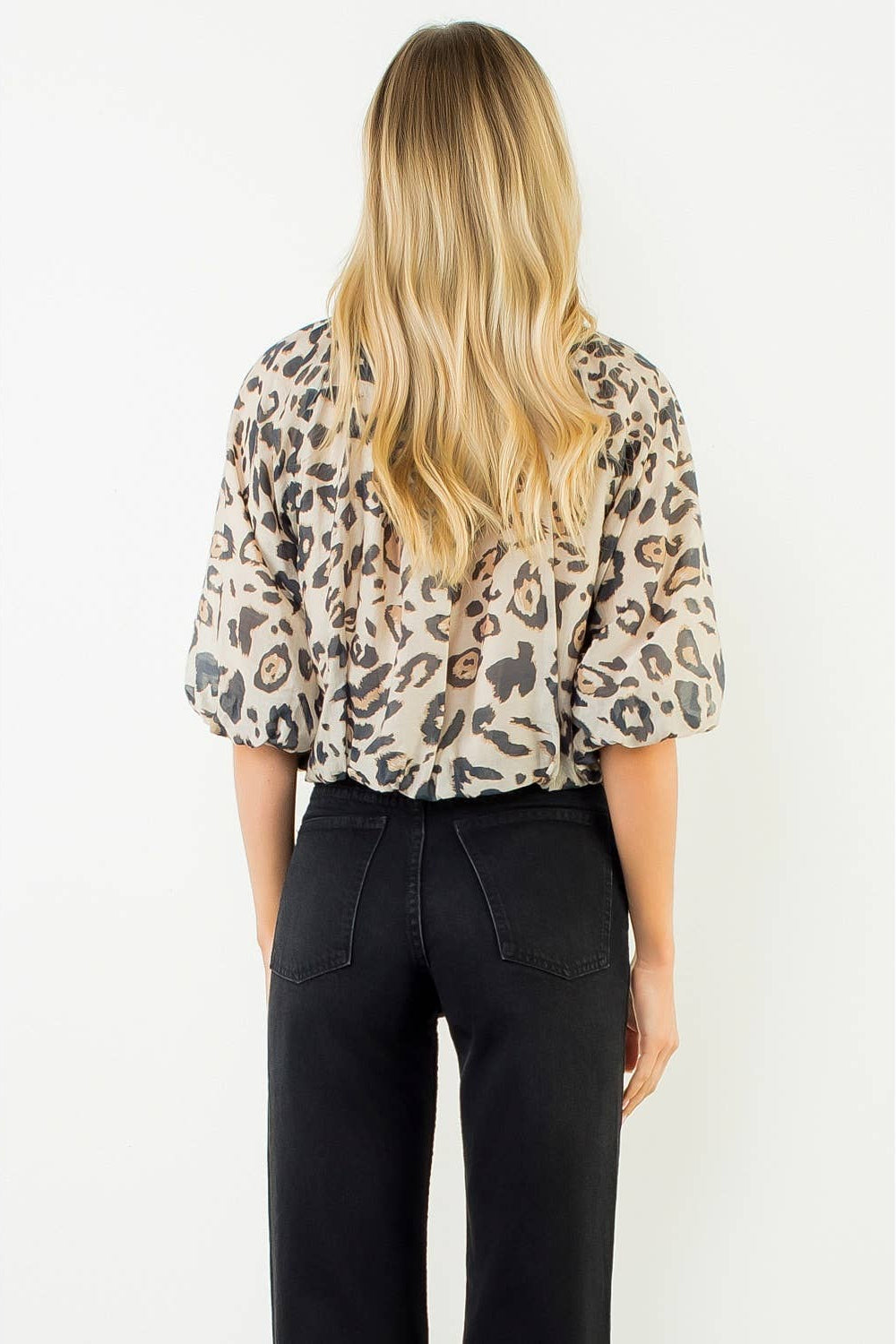 A woman viewed from behind wearing a leopard-print short-sleeve blouse with puff sleeves and black pants, standing against a plain white background.