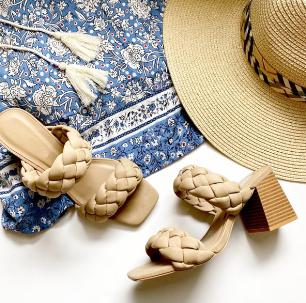A pair of beige braided heel sandals displayed alongside a blue patterned fabric and a straw hat.