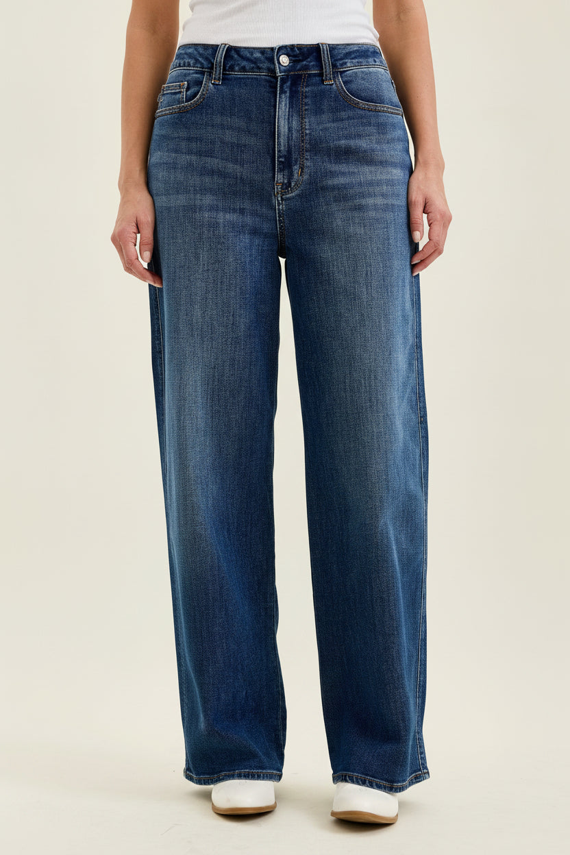 A person wearing dark blue wide-leg jeans with a white top and white shoes, shown from the waist down against a plain light beige studio background.