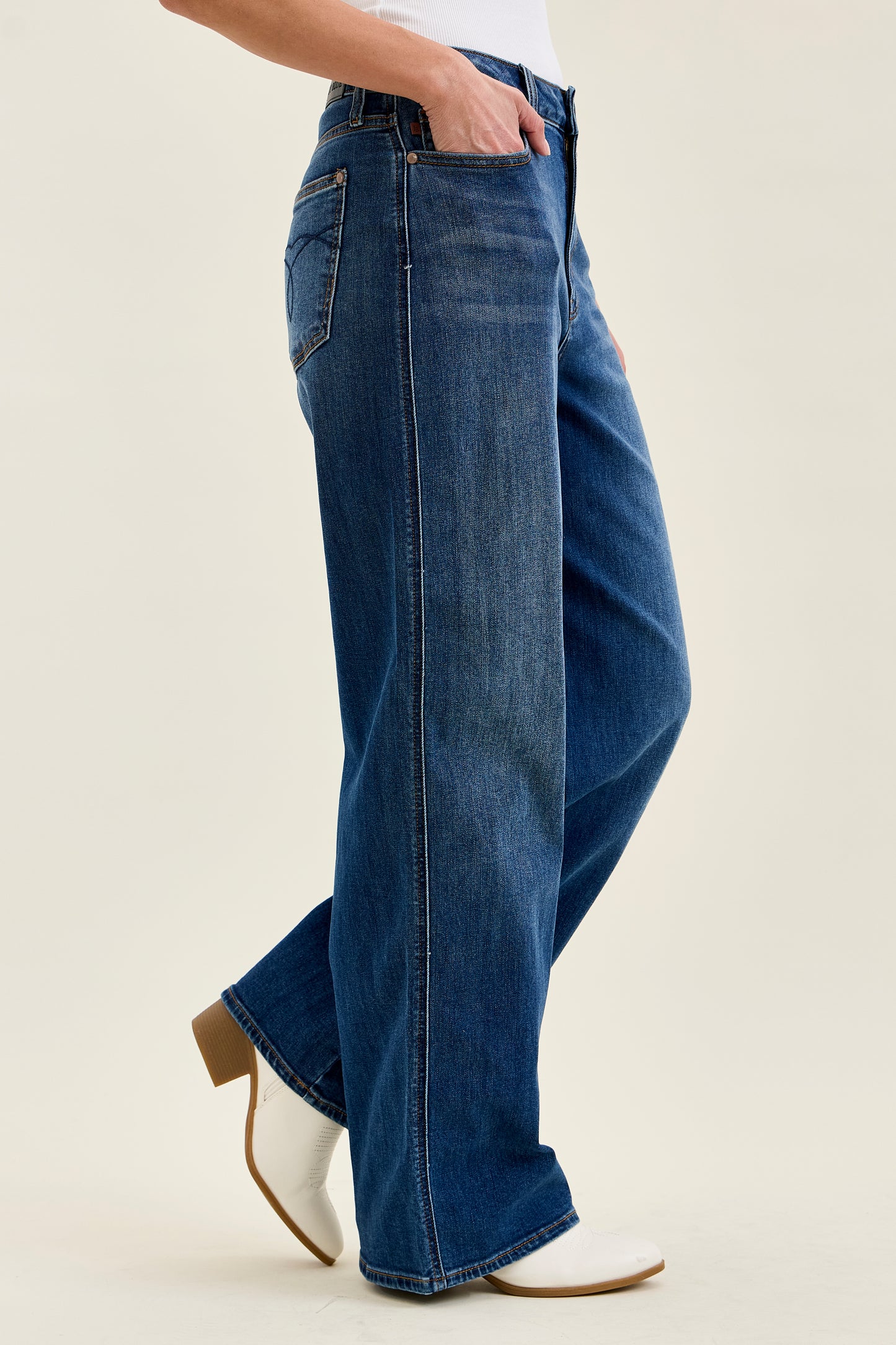 A person wearing blue wide-leg jeans with a white top and white ankle boots, shown from the waist down against a plain light beige studio background.