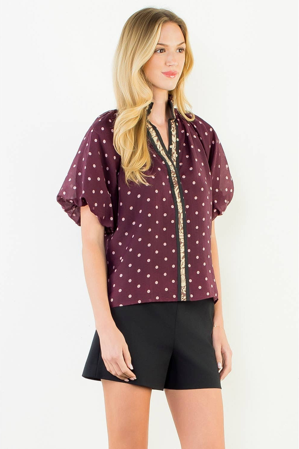A woman modeling a short-sleeve button-up blouse in burgundy with white polka dots and a gold trim down the front, paired with black shorts, against a plain white background.