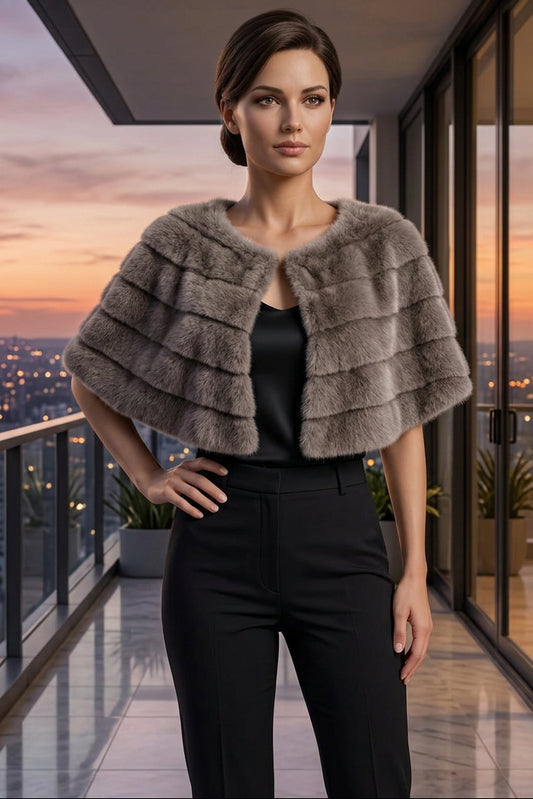 A woman wearing a plush, tiered taupe-brown faux fur cape jacket over a white top and white trousers, standing on a modern balcony with a city skyline in the background.