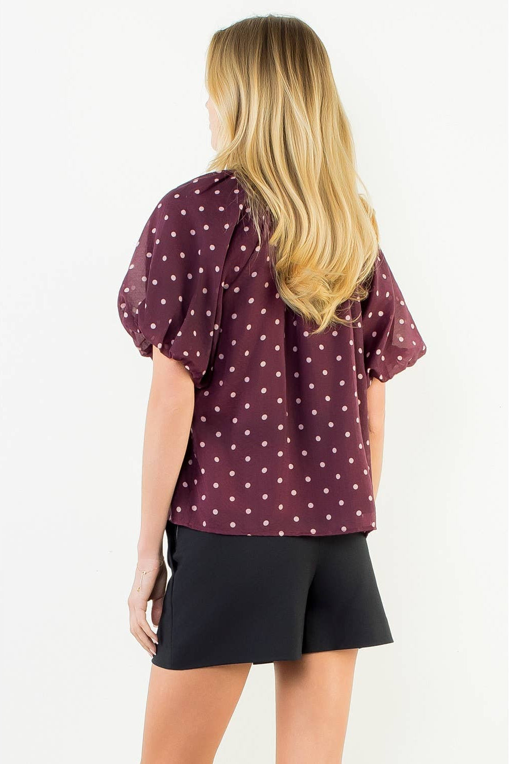 A woman shown from the back wearing a short-sleeve burgundy top with a white polka dot pattern and black shorts, set against a plain white studio background.