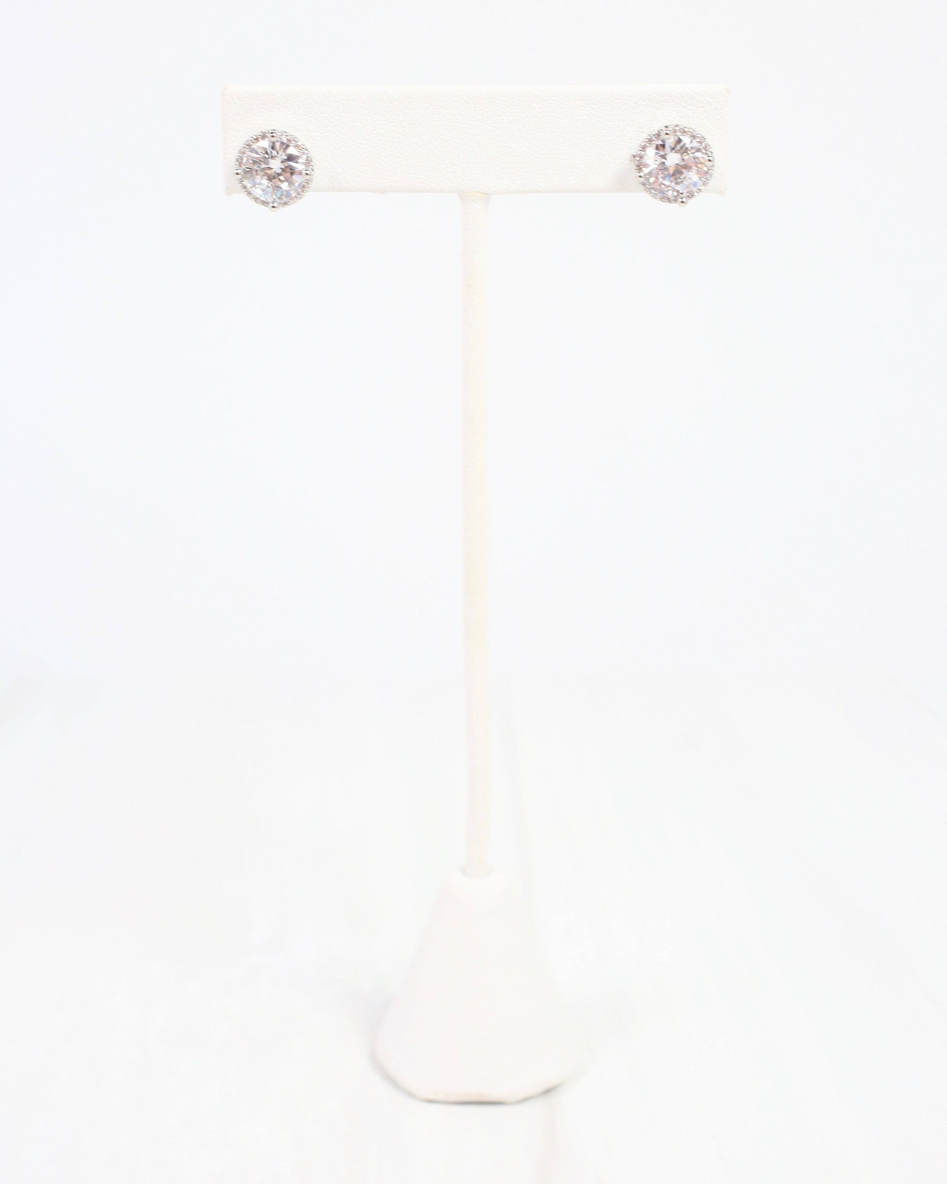 A pair of round stud earrings with sparkling clear gemstones in a silver-toned setting, displayed on a white jewelry stand against a white background.