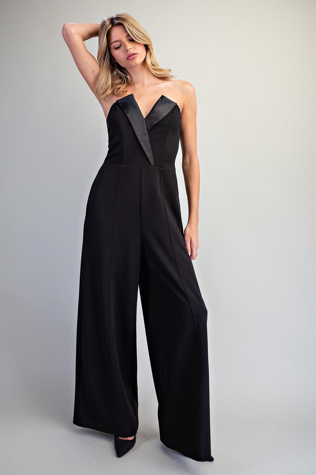 A woman modeling a black strapless wide-leg jumpsuit with contrasting black satin-like trim at the sweetheart neckline, photographed against a plain light gray studio background.