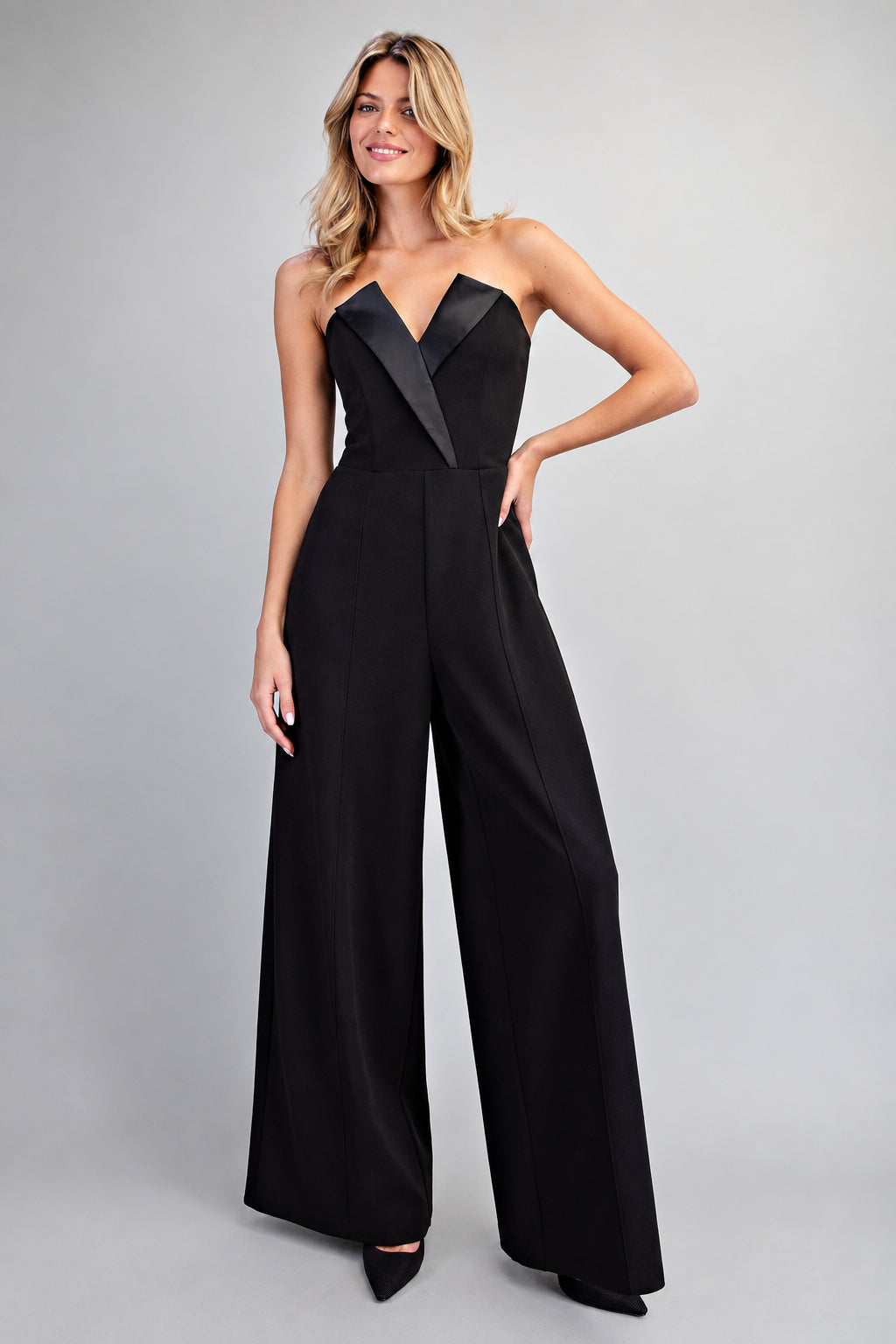 A woman modeling a black strapless wide-leg jumpsuit with contrasting black satin lapels, photographed against a neutral light gray studio background.