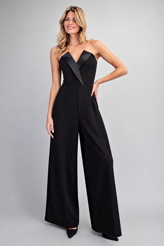 A woman modeling a black strapless wide-leg jumpsuit with contrasting black satin lapels, photographed against a neutral light gray studio background.