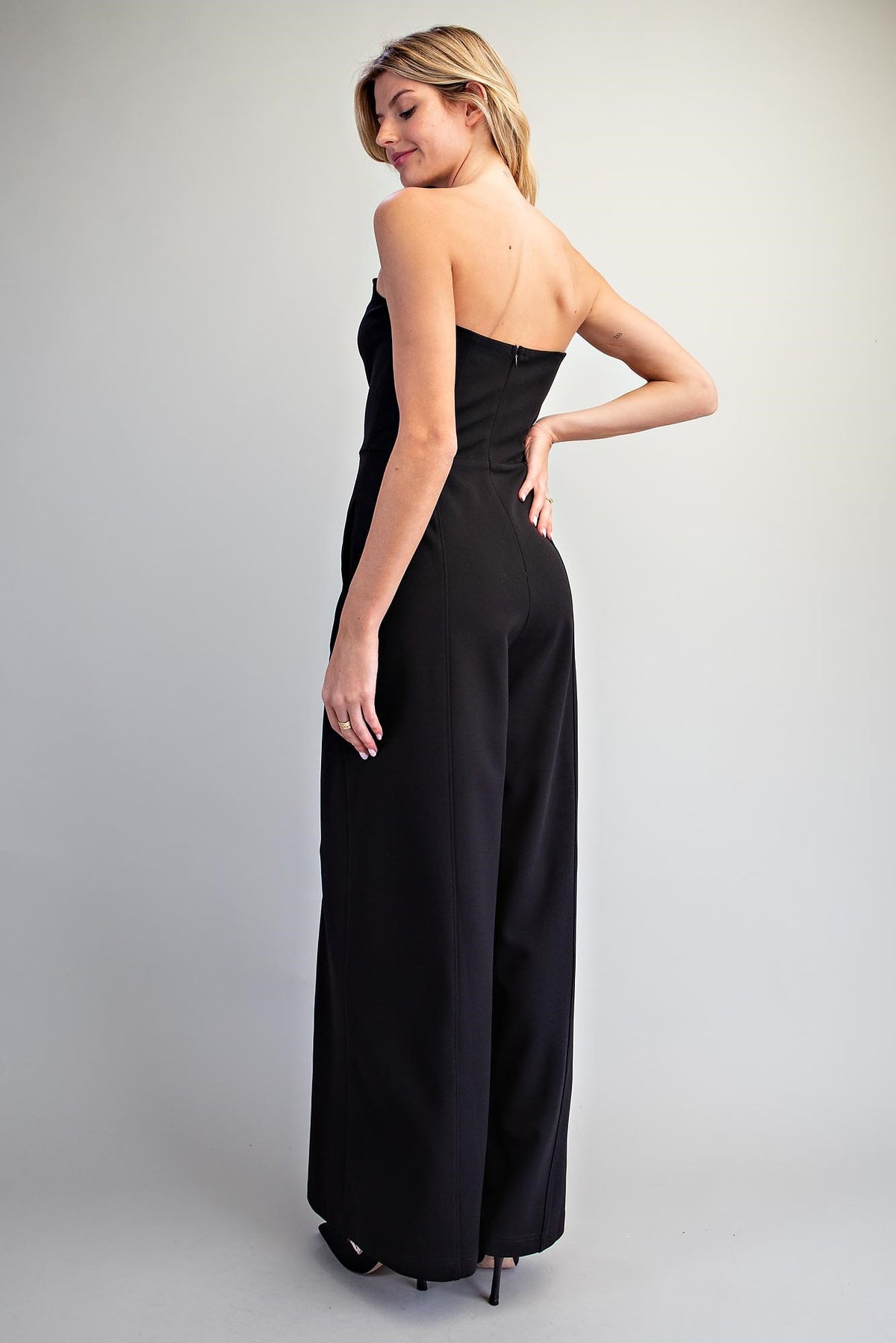 A woman modeling a black strapless maxi jumpsuit with wide legs, shown from a three-quarters back view against a light gray studio background.