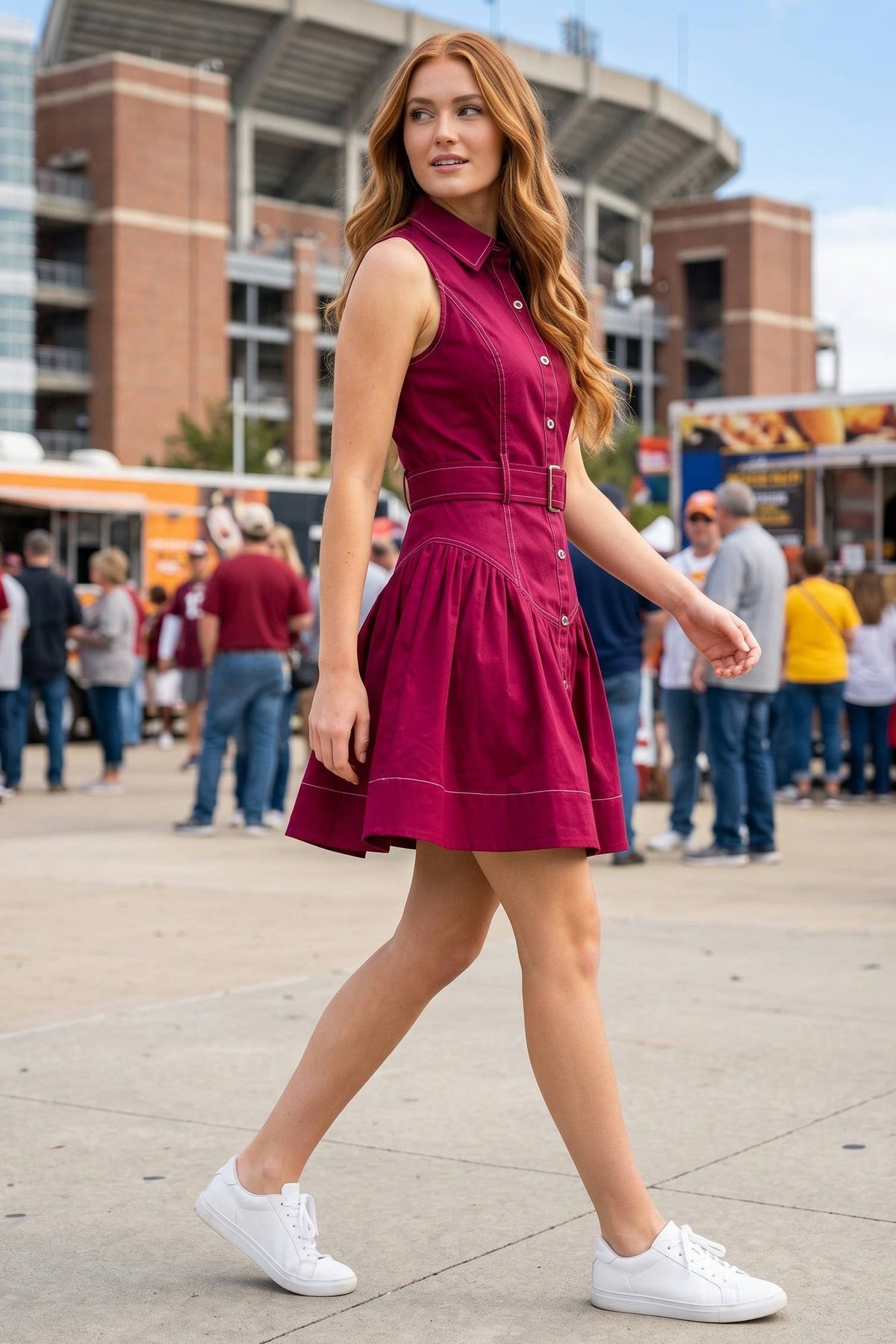 A woman with long wavy red hair walks outdoors in front of a stadium, wearing a sleeveless garnet
 mini dress with white sneakers.