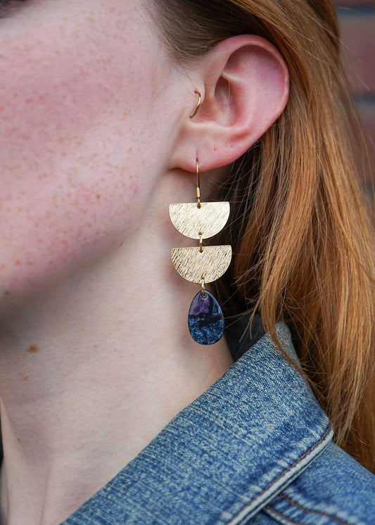 Close-up of a person wearing gold earrings with purple stones, against a blurred background.