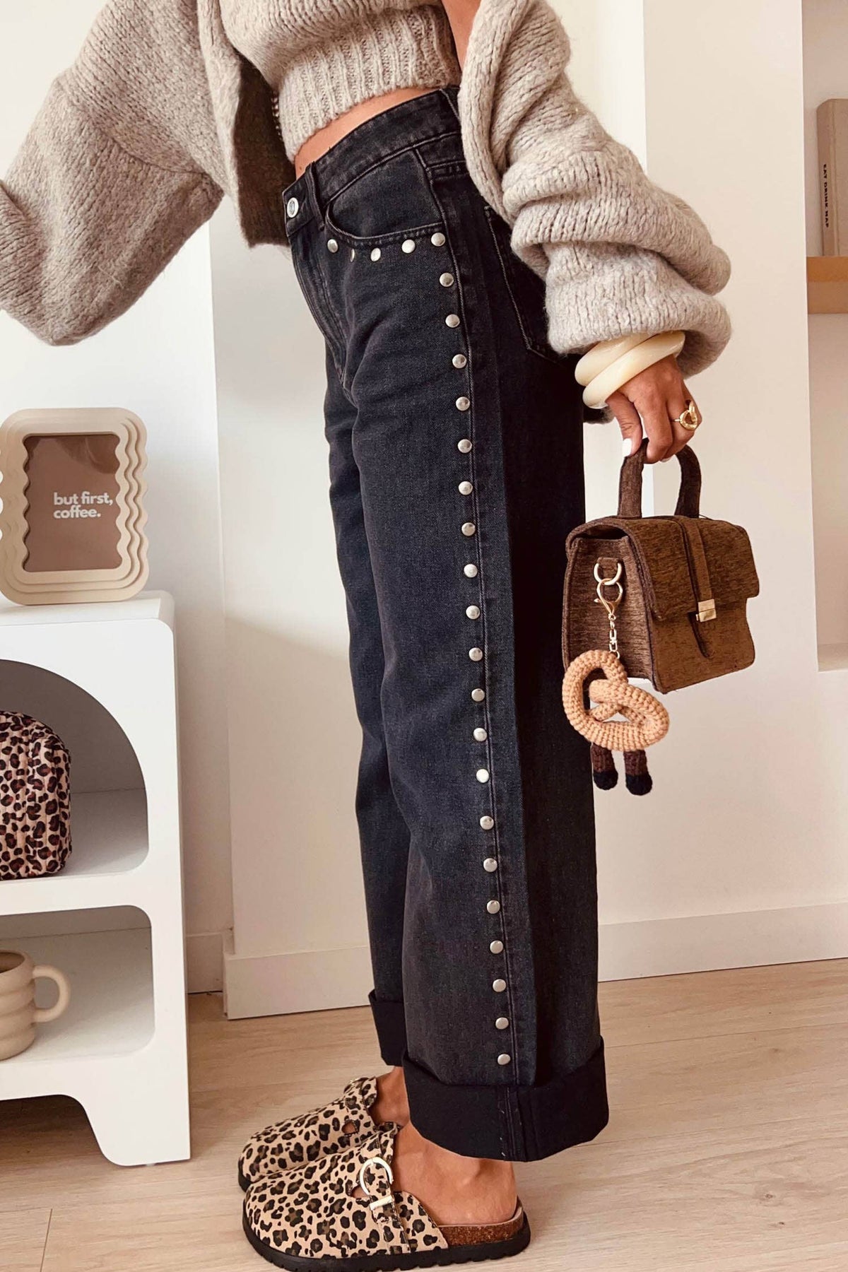 Person wearing a beige sweater, dark jeans with button details, and leopard print slippers holding a brown handbag.