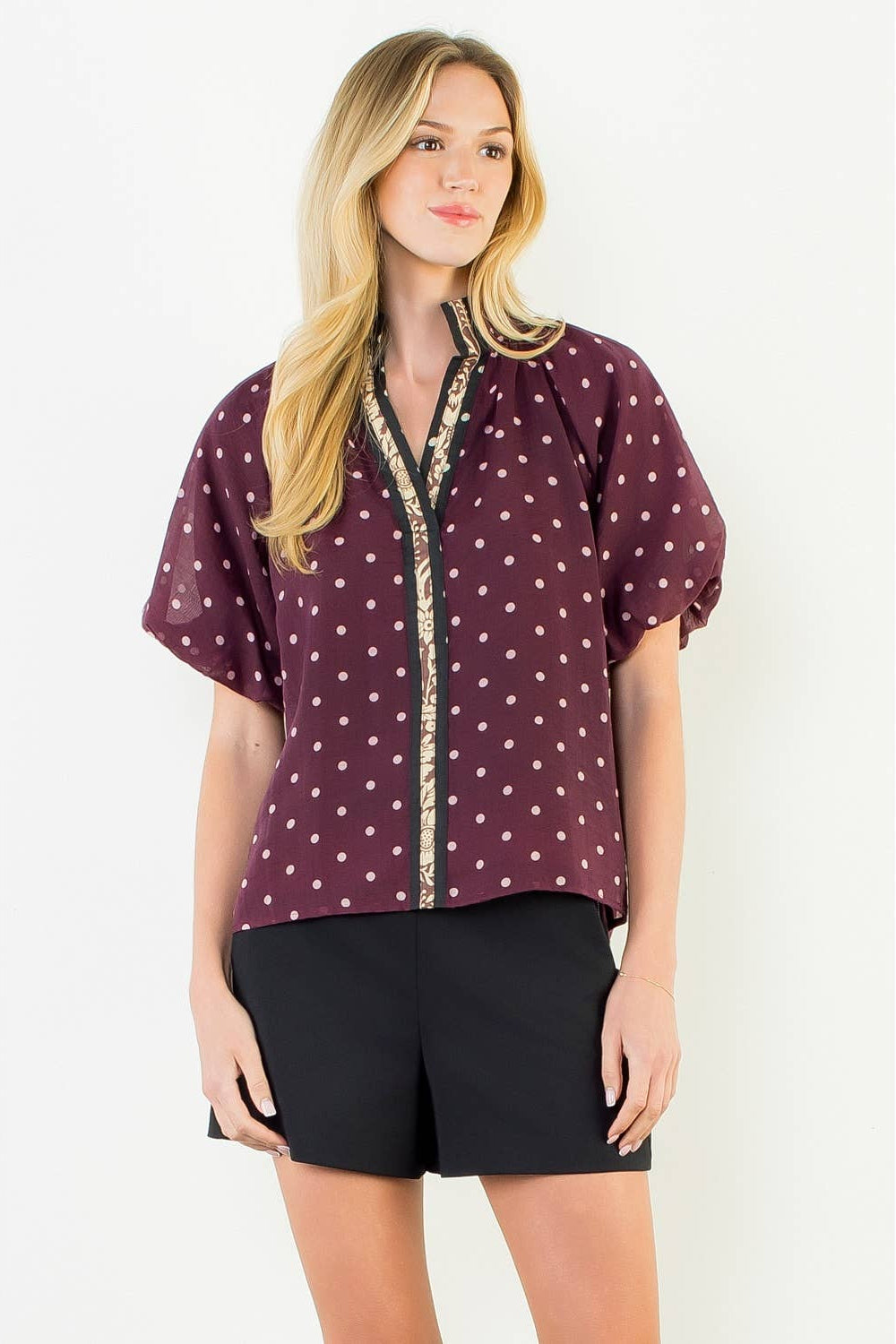 A woman modeling a short-sleeve button-up blouse in burgundy with a white polka dot pattern and a gold-trimmed V-neckline, paired with black shorts against a plain white studio background.