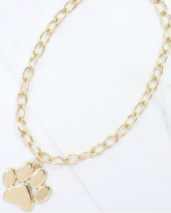 A gold chain necklace with a polished paw print pendant, displayed on a white marble surface.