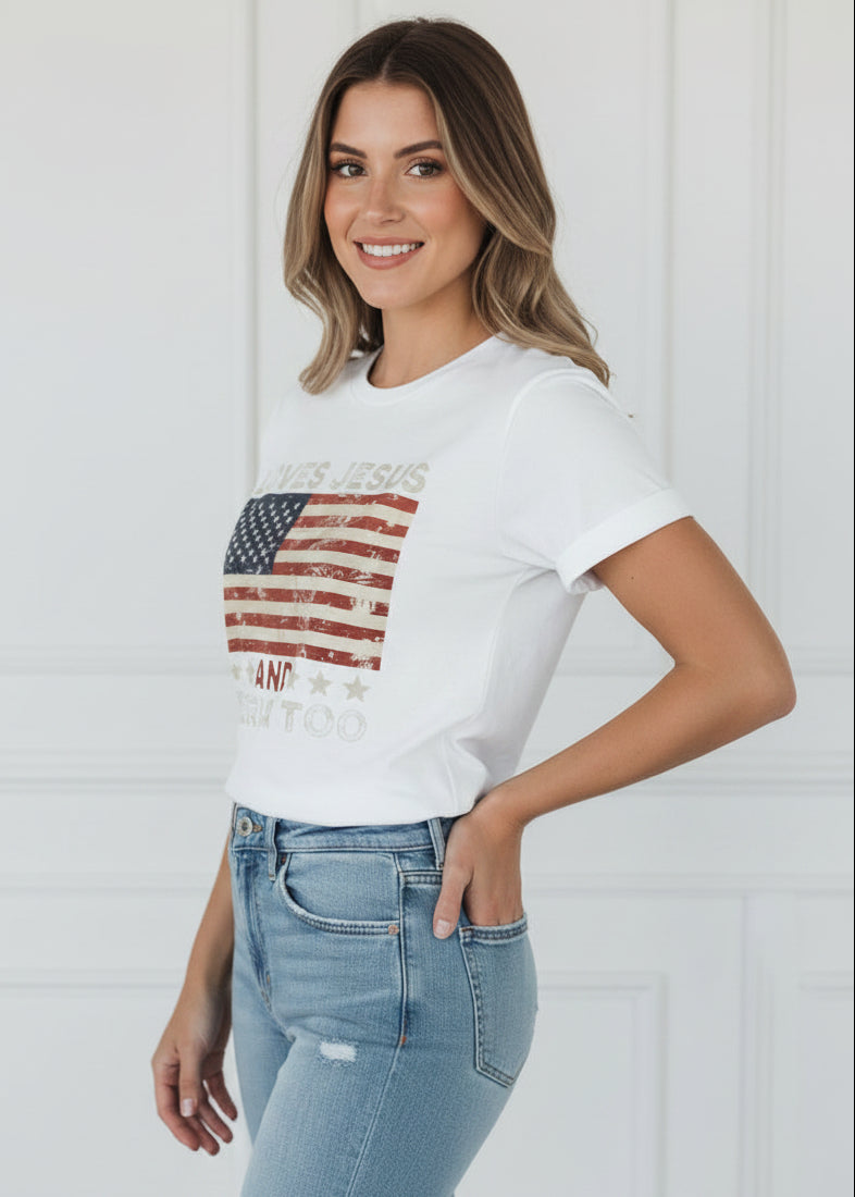 White t-shirt with American flag design and text 'LOVES JESUS AND AMERICA TOO' on a wooden background.