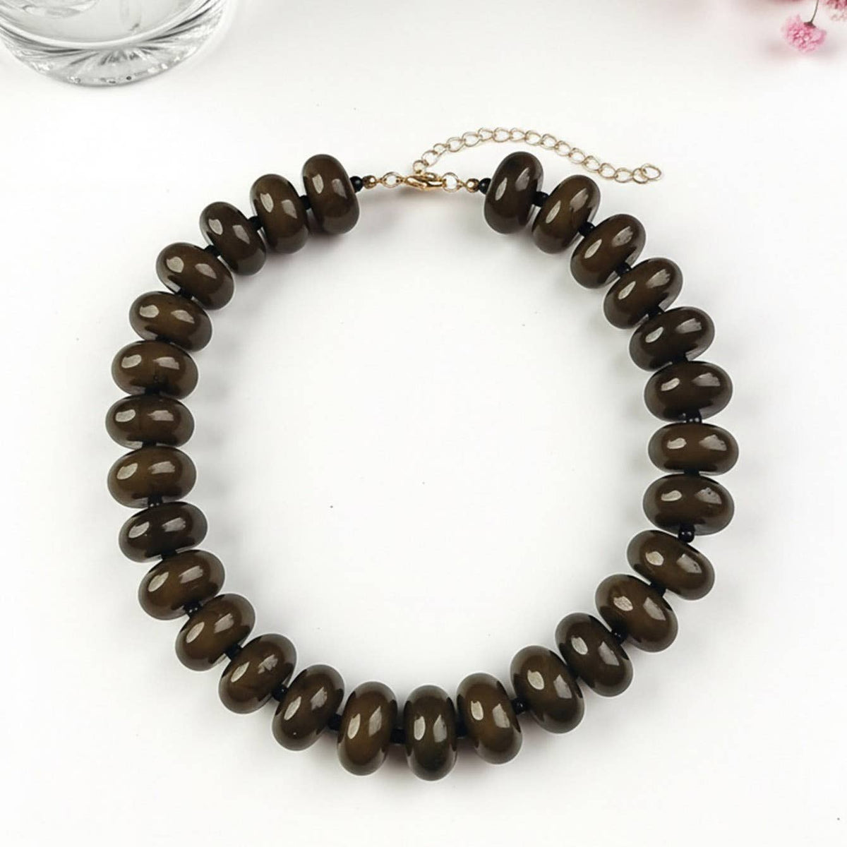 A dark brown glossy beaded necklace with a gold-tone chain extender, displayed on a clean white surface with decorative pink flowers and a glass in the background.