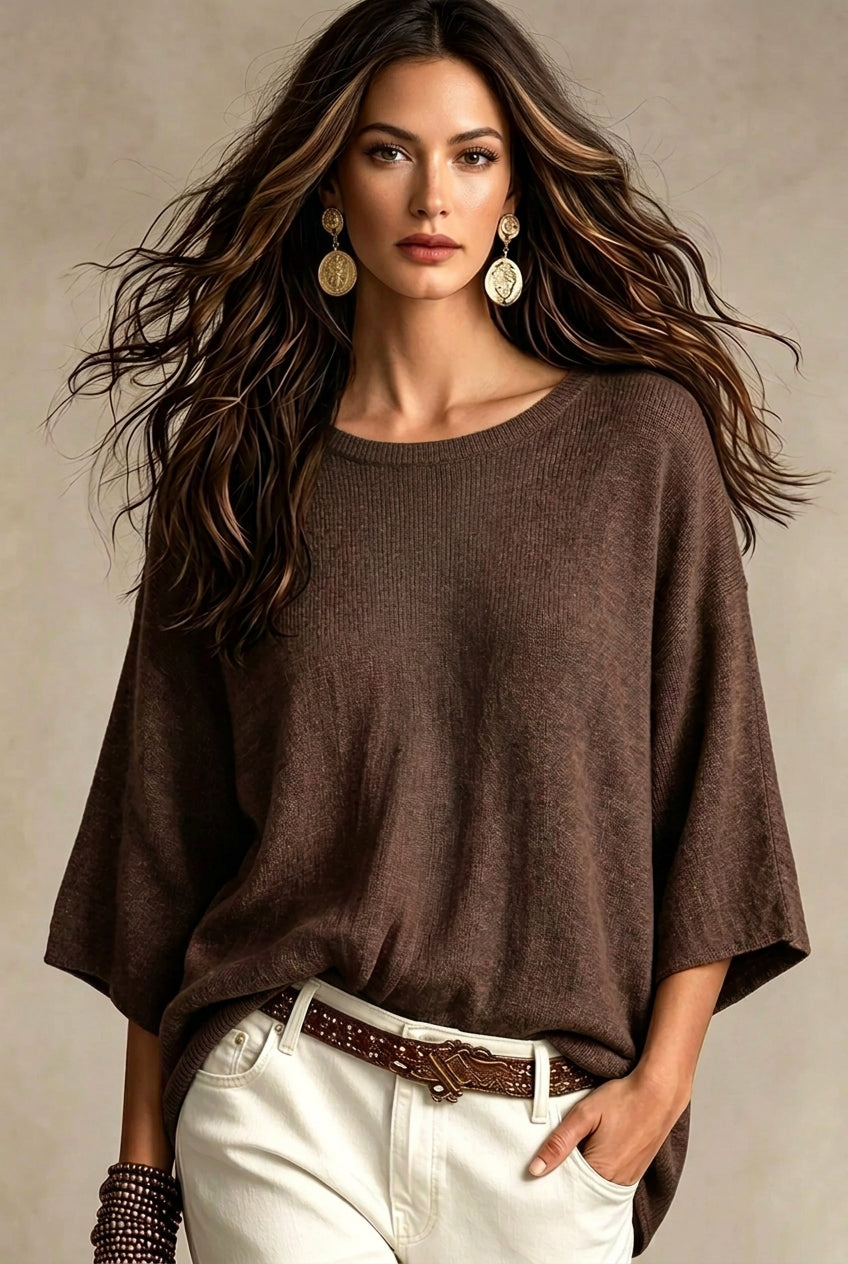 A woman wearing a brown short-sleeve knit top with wide batwing sleeves, styled with layered gold necklaces, white pants, and a brown belt against a neutral beige background.