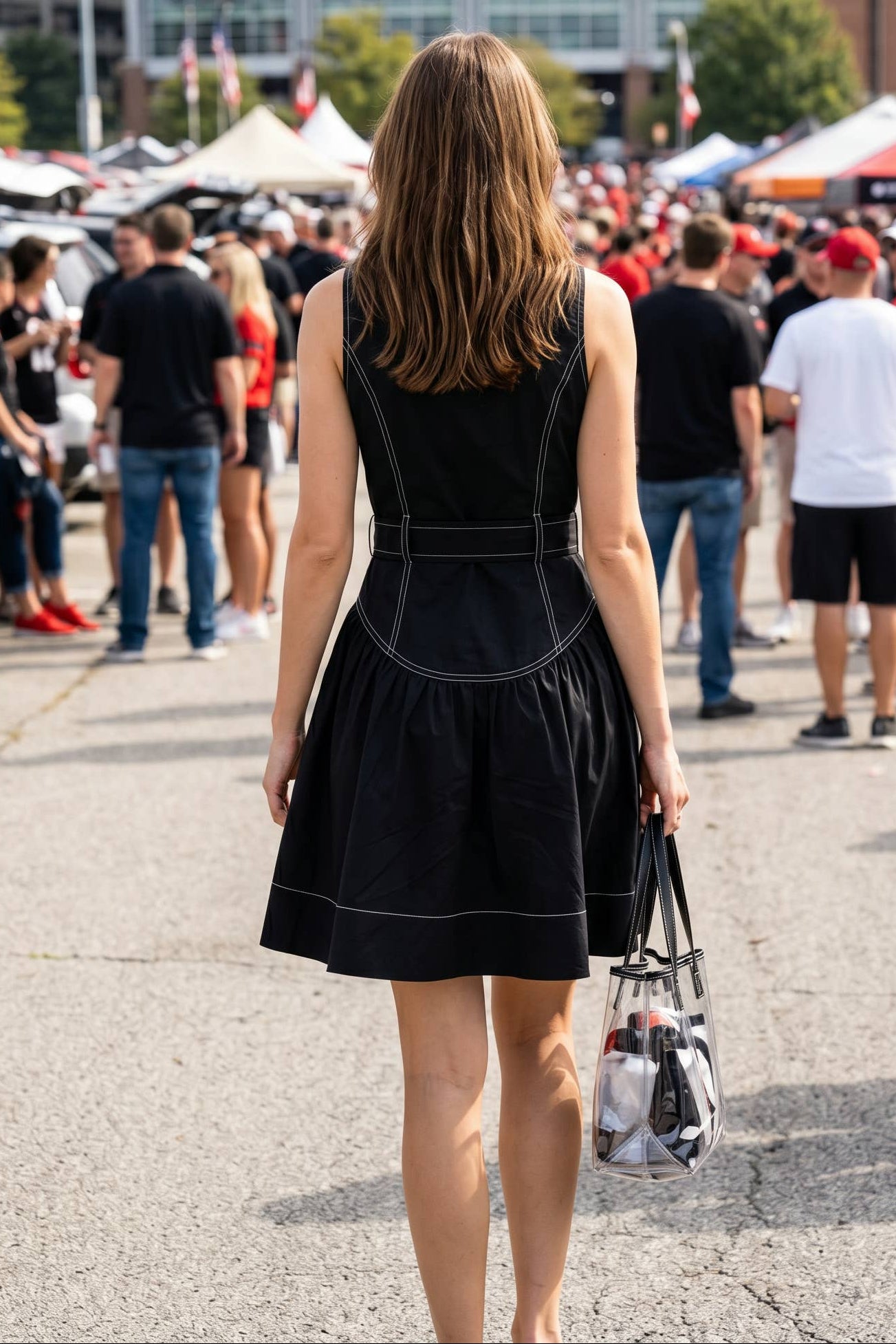 A woman viewed from behind wearing a black sleeveless mini dress with white contrast piping, standing in a crowded outdoor stadium parking area.