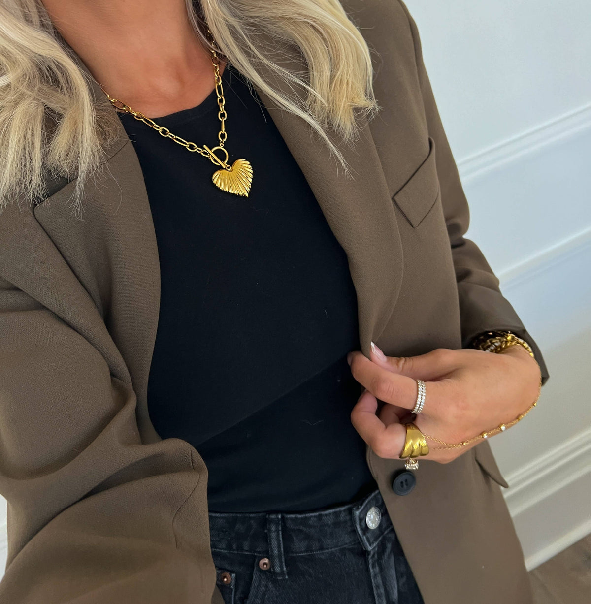 A close-up lifestyle shot of a woman wearing a gold heart-shaped pendant necklace over a black top and brown blazer.
