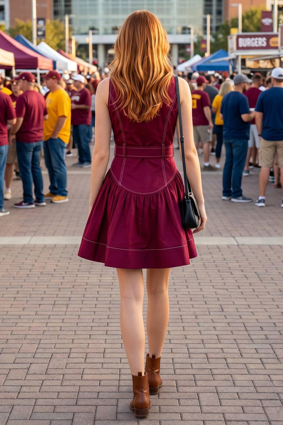 A woman with long red hair is seen from behind walking on a brick plaza toward a stadium, wearing a sleeveless burgundy mini dress with white piping and brown ankle boots, while carrying a black handbag.