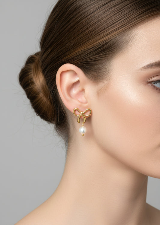 Gold bow-shaped earrings with pearl drops held in a hand against a neutral background