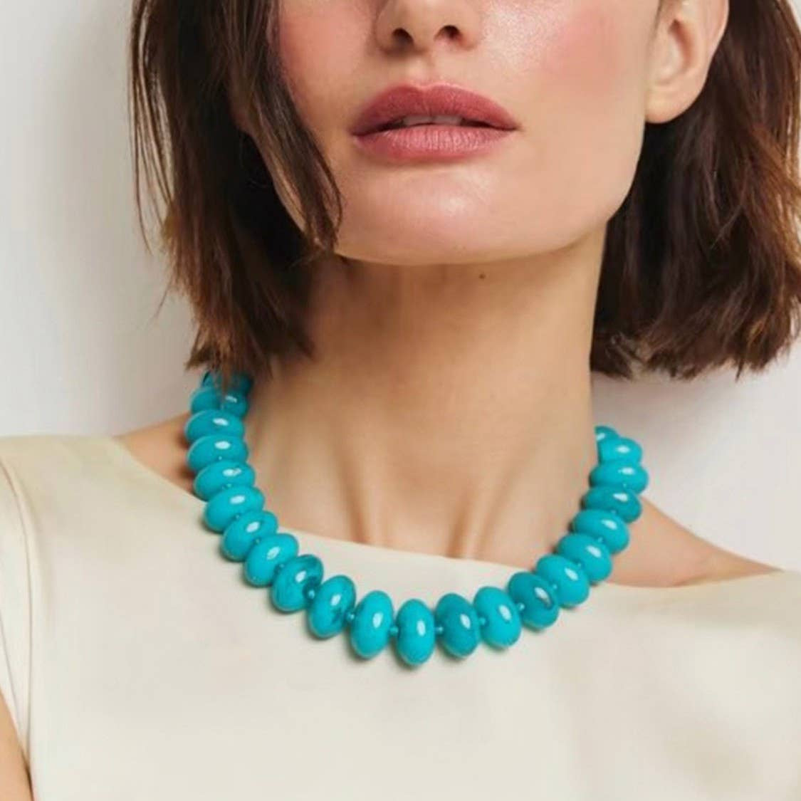 A close-up of a model wearing a chunky turquoise beaded necklace with large, glossy oval beads, paired with a cream-colored top.