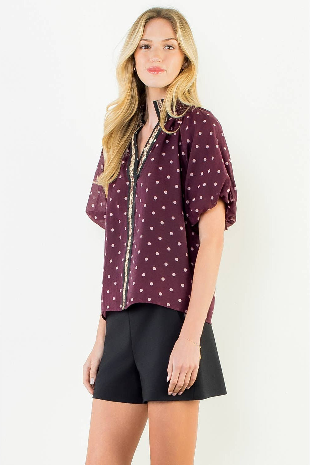 A woman modeling a short-sleeve button-up blouse in burgundy with a white polka dot pattern and a gold trim down the front, paired with black shorts, against a plain white background.