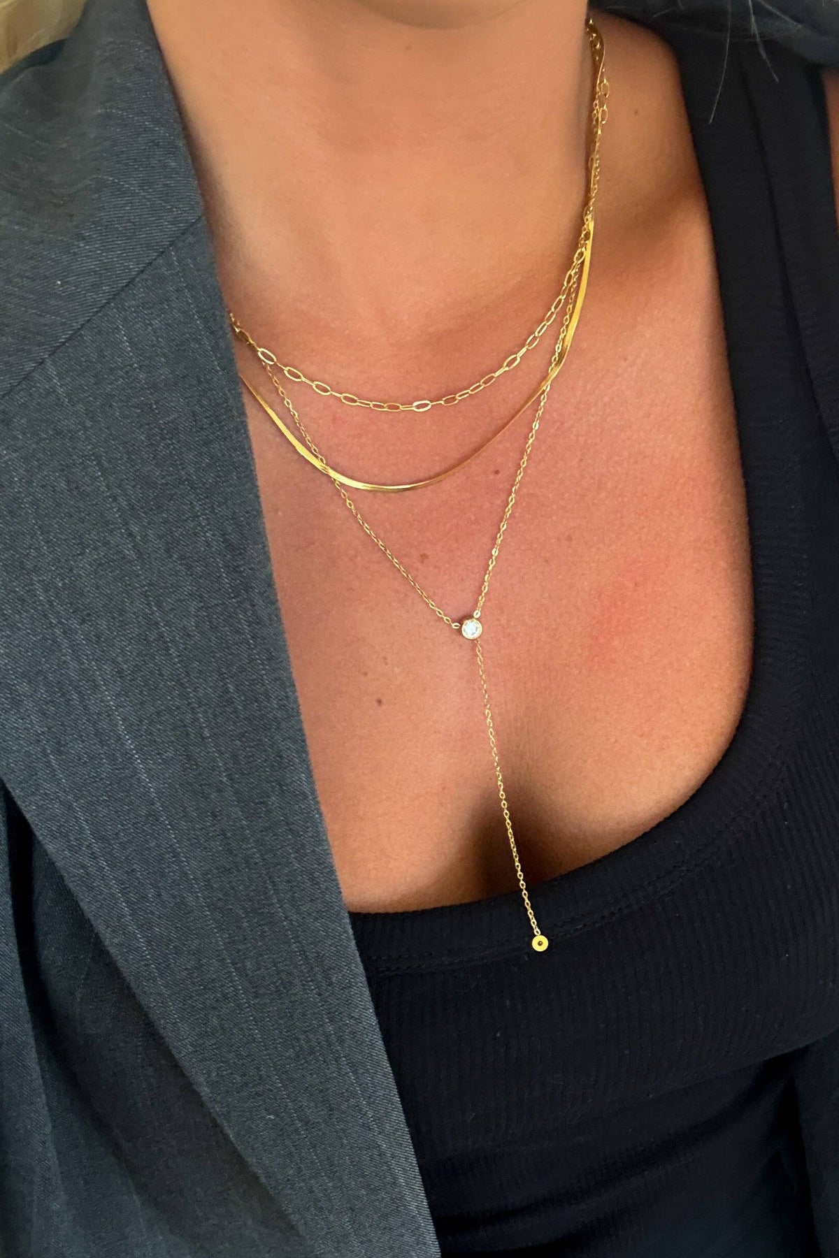 A close-up of a person wearing layered gold necklaces over a black ribbed tank top and a gray blazer.