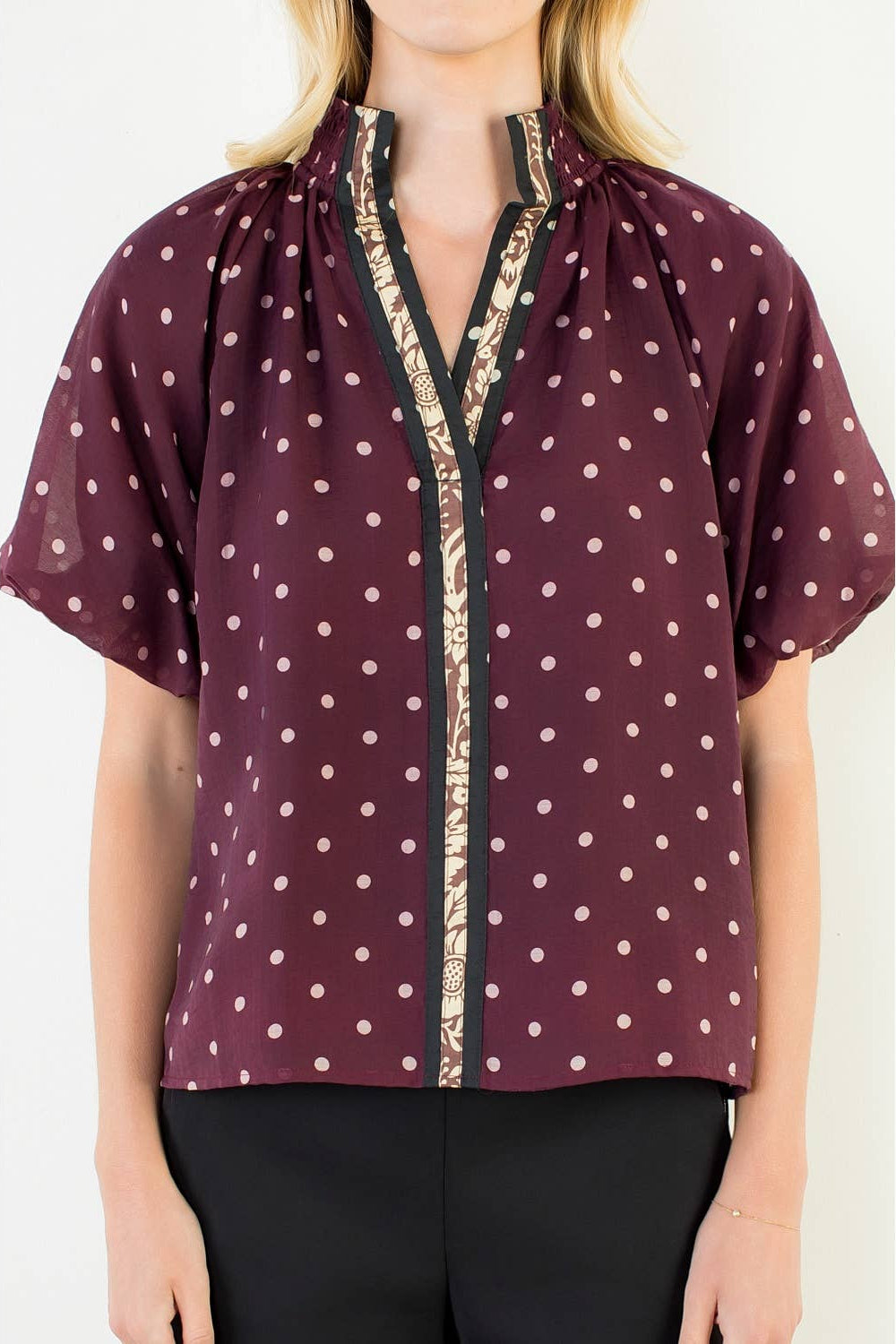A woman wearing a short-sleeve blouse with a burgundy base, white polka dots, and a beige patterned trim along the split neckline and front placket, paired with black pants.