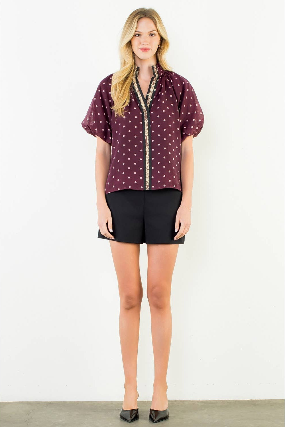 A woman stands against a plain white wall wearing a short-sleeve burgundy blouse with small white polka dots and a gold trim down the front, paired with black shorts and black pointed heels.