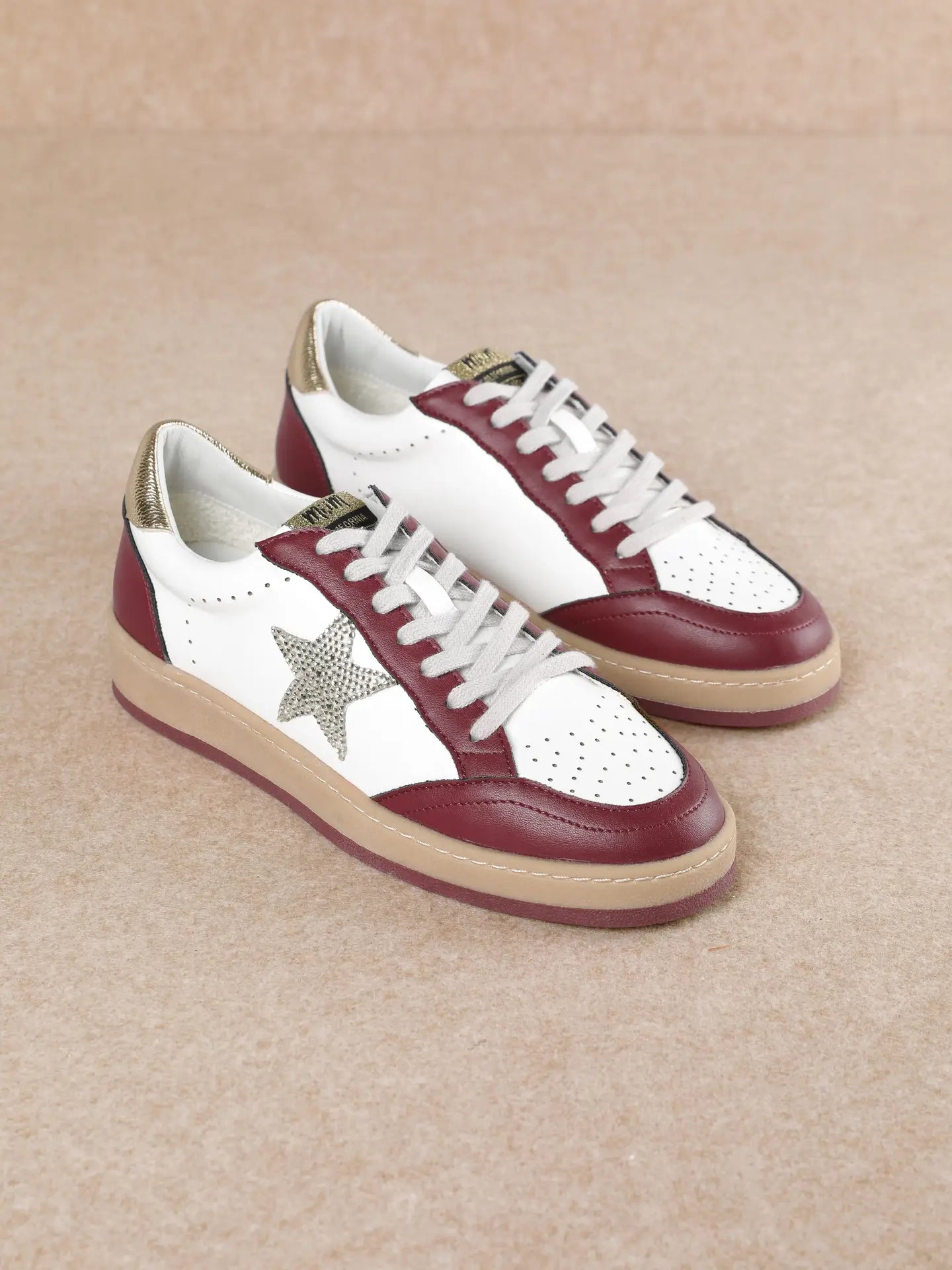 A pair of low-top sneakers with white leather panels, burgundy leather overlays, a metallic silver glitter star on the side, white laces, and beige soles, set against a neutral beige background.