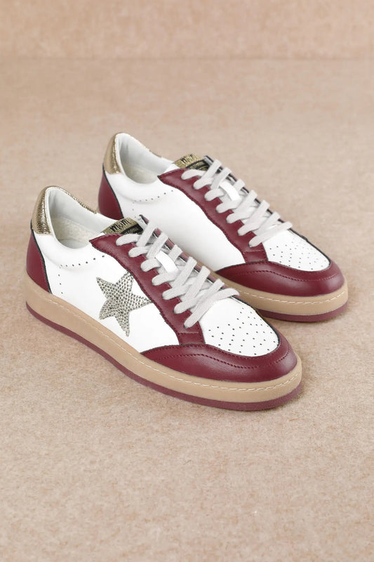 A pair of low-top sneakers with white leather panels, burgundy leather overlays, a metallic silver glitter star on the side, white laces, and beige soles, set against a neutral beige background.
