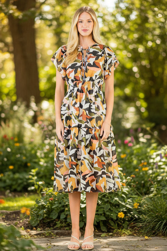 A woman modeling a short-sleeve midi dress with a colorful abstract print in orange, yellow, green, black, and white, standing against a plain white wall.