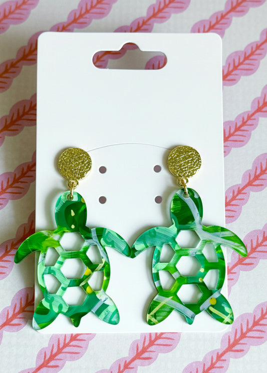 Green turtle-shaped earrings on a white card with 'SR' branding, against a pink and white striped background.