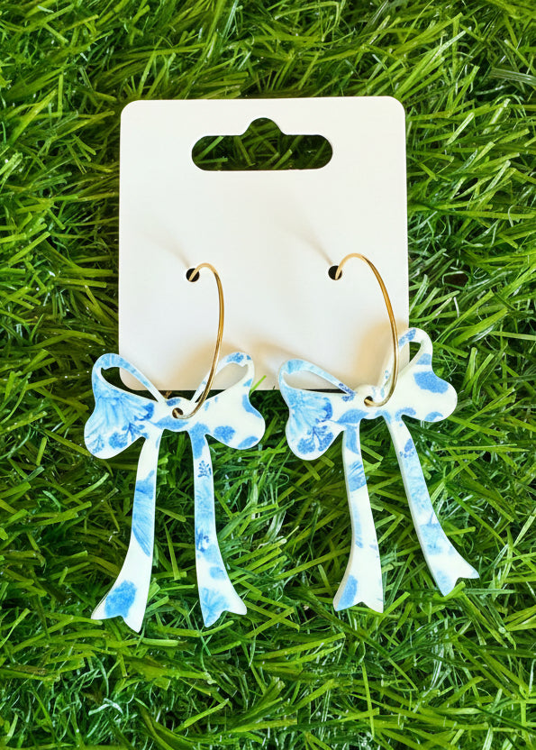 Pair of earrings with blue and white designs on a pink card with 'SR' branding, placed on green grass.