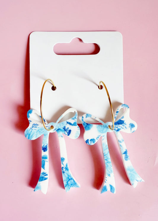 Pair of earrings with blue floral design on a white card with 'SH' logo against a pink background