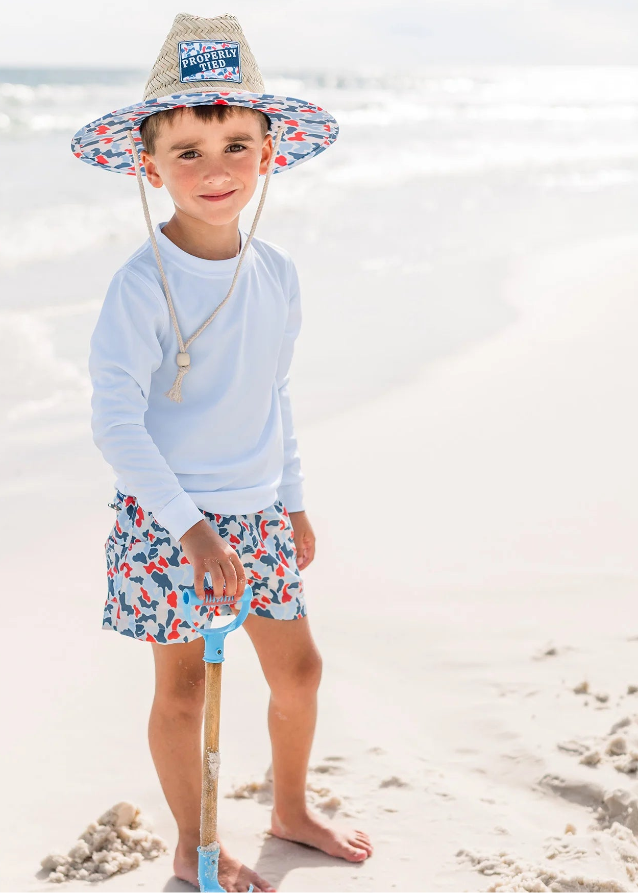 Child on a beach wearing a sun hat, light blue shirt, and colorful shorts with a shovel.