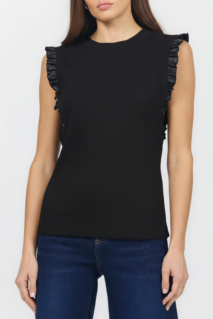 A woman wearing a sleeveless black top with ruffled trim along the shoulders and sides, paired with dark blue jeans, against a plain light gray studio background.