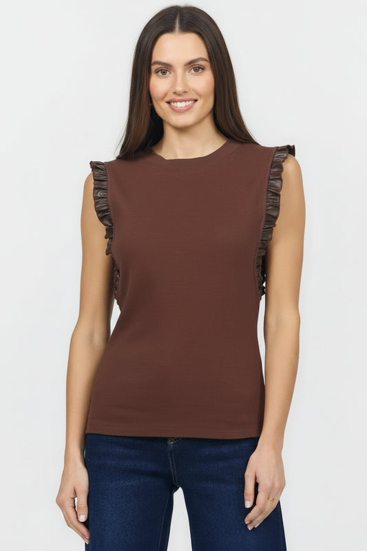A woman wearing a sleeveless brown top with dark ruffle trim along the shoulders, paired with dark blue jeans, against a light gray studio background.