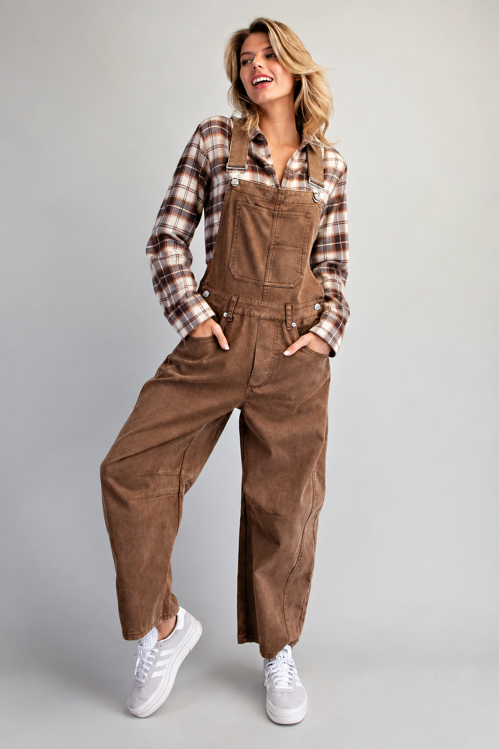 A woman modeling brown corduroy bib overalls over a brown and beige plaid flannel shirt, paired with white sneakers, against a solid gray studio background.