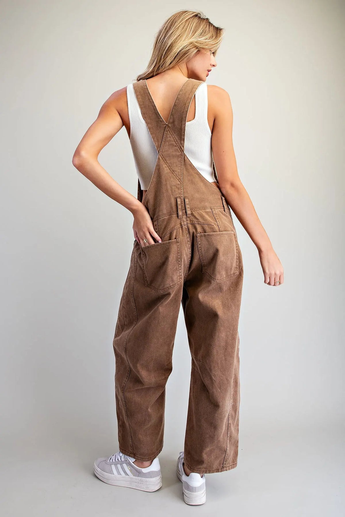 A woman modeling brown corduroy bib overalls with wide legs, shown from a back-three-quarter view. She is wearing a white tank top and white sneakers against a light gray studio background.